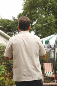 A man viewed from behind wearing a light beige short-sleeve linen shirt and dark pants, standing in an outdoor backyard setting with trees, a patio umbrella, and a chair in the background.