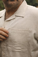 Close-up of a man wearing a light beige linen short-sleeve button-up shirt with a chest pocket, showing the fabric texture and collar.