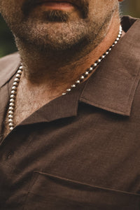 A close-up shot of a man wearing a silver beaded necklace over a brown collared shirt.
