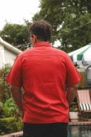 A man viewed from behind wearing a bright red short-sleeve button-up shirt and dark pants, standing outdoors near a pool with trees and patio furniture in the background.
