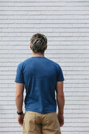 Man with short blond hair wearing blue t-shirt and beige pants standing against white brick wall