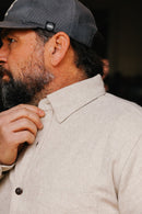 Bearded man adjusting collar of cream flannel Oxford shirt wearing dark cap