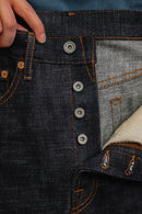Close-up of hand holding dark indigo raw denim jeans with orange stitching and metal button fly